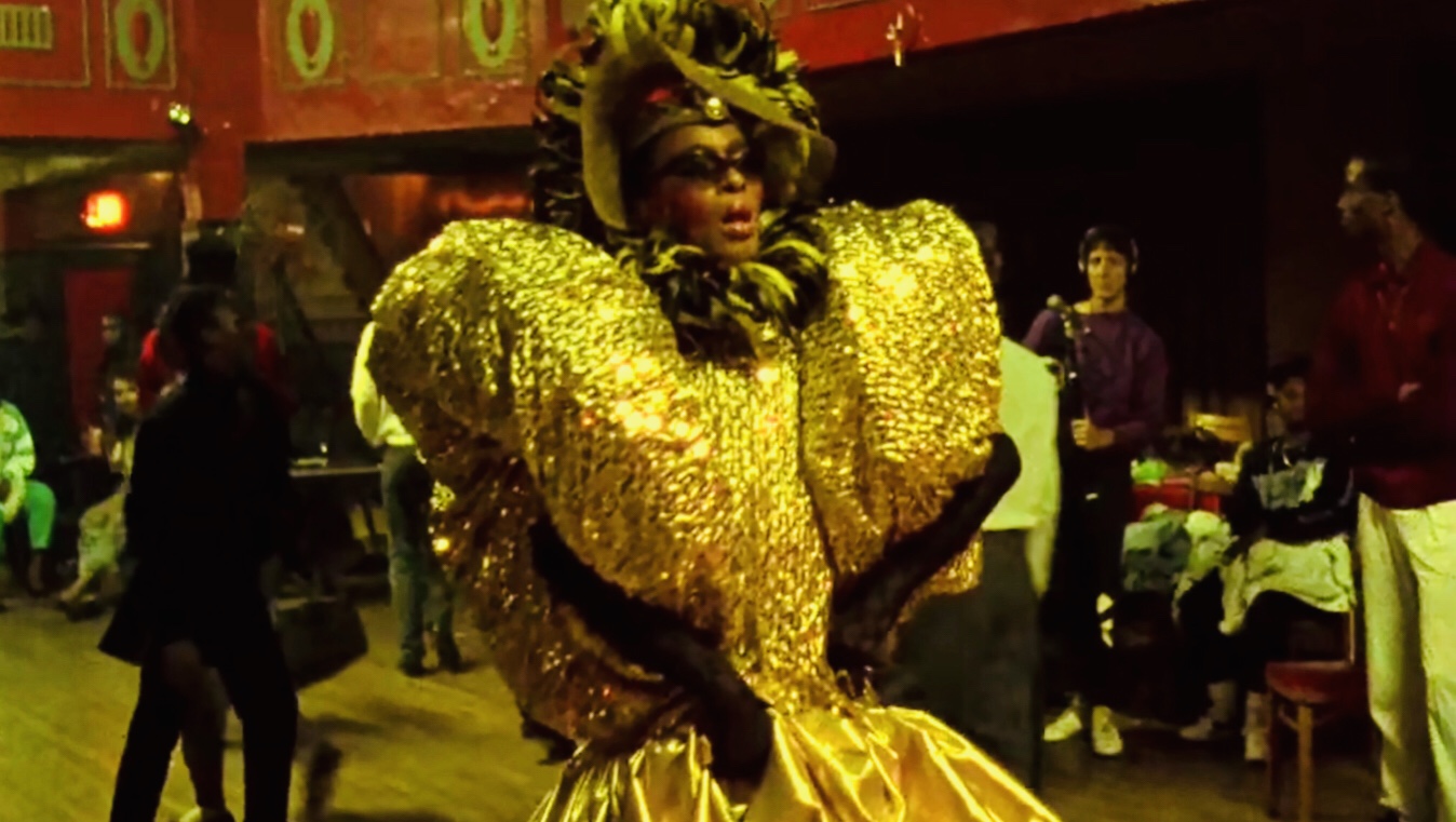 Image is from the film 'Paris is Burning' (1990). A drag queen walks in an extravagant gold gown in a ball. The gown is metallic and has big poofy sleeves.